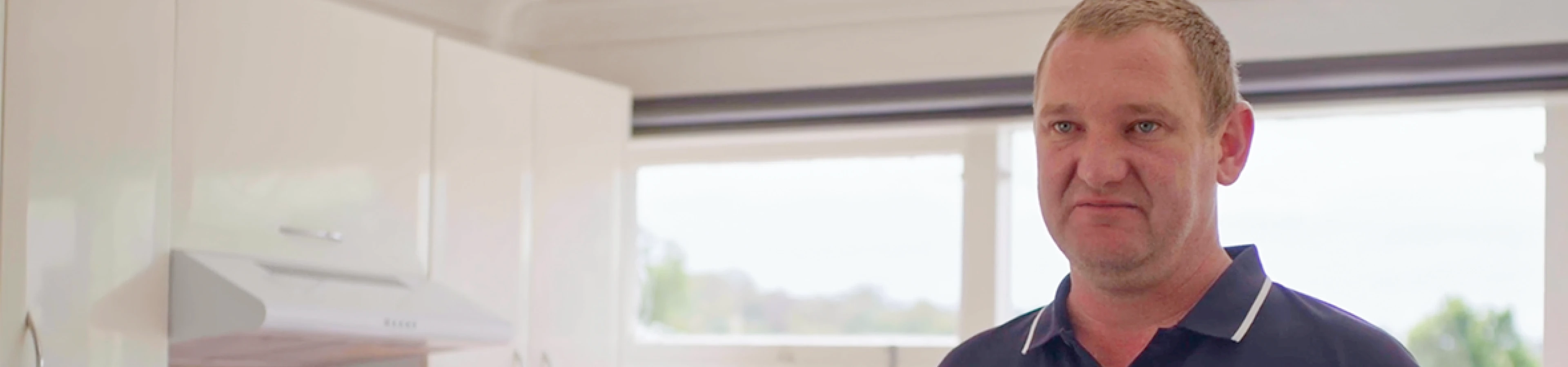 a man wearing a dark navy polo shirt standing inside a bright kitchen.