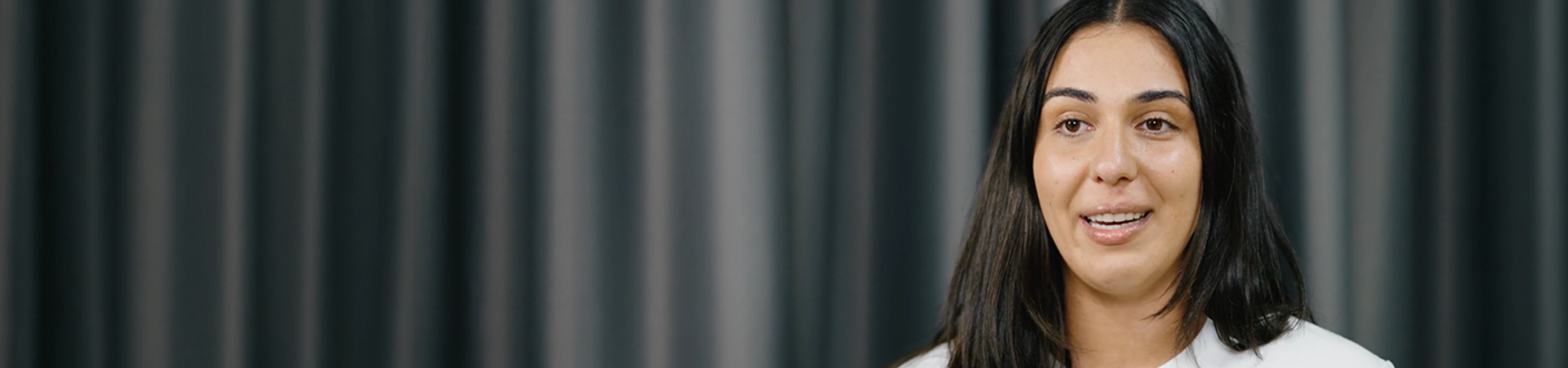 A woman with dark hair speaking against a gray curtain backdrop.