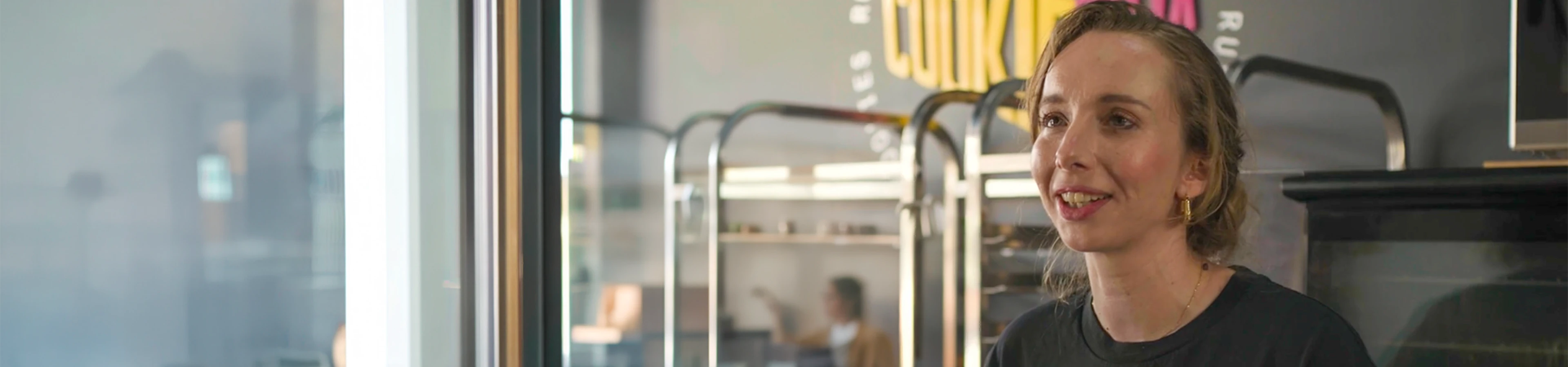 A close-up portrait of a woman smiling and speaking in front of commercial baking racks inside a kitchen or bakery setting.